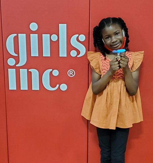 a little girl stands beside a girls inc logo in a orange shirt and black pants